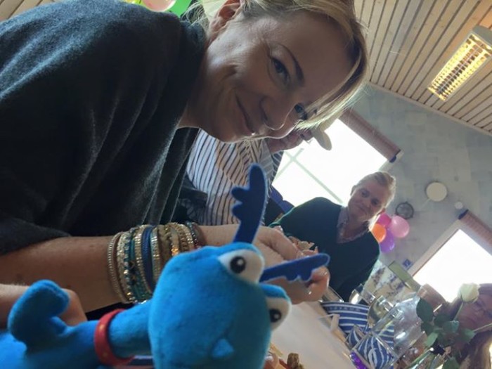 En kvinna sitter leende vid ett bord som är dukat för middag. Med henne sitter funkibators maskot-den blå dinosaurien.