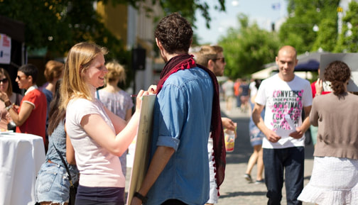 Flera unga människor är samlade ute i en stad. Närmast kameran är en kille som står i centrum och en tjej som fäster en affisch på hans bröst.