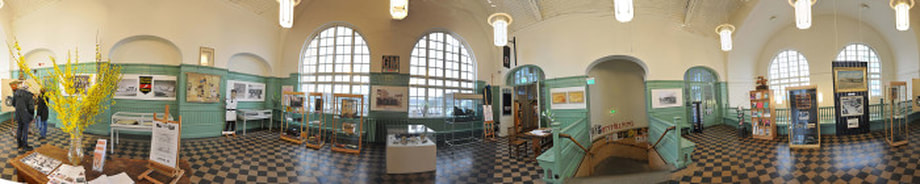 En panoramabild på Alvesta utställningshall. Golvet är rutigt och på de turkosa väggarna hänger tavlor.