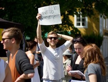 En ung kille håller en affisch högt upp i luften. Runt honom står andra unga människor, både tjejer och killar. Affischens text är: Vad vill du förändra?