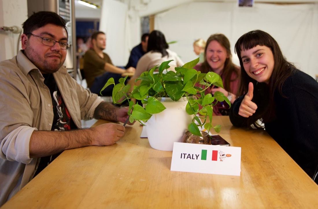 Vid ett bord i ett rum sitter några unga människor. På bordet står en bild av italiensk flagga och en växtkruka. Alla tittar in i kameran. Bakom dem sitter andra människor.